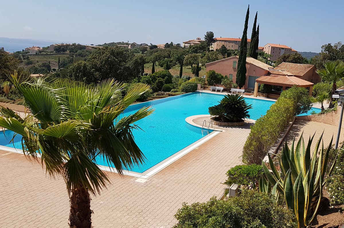 Großer Außenpool mit azurblauem Wasser, umgeben von Palmen, Agaven und mediterranen Gebäuden unter klarem Himmel an der Côte d’Azur.
